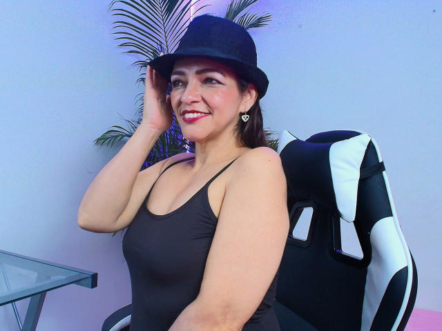 CarolMissx with dark skin, medium tits, brown eyes, wearing a black tank top and black hat, smiling, hand on hat, sitting in a black and white gaming chair.