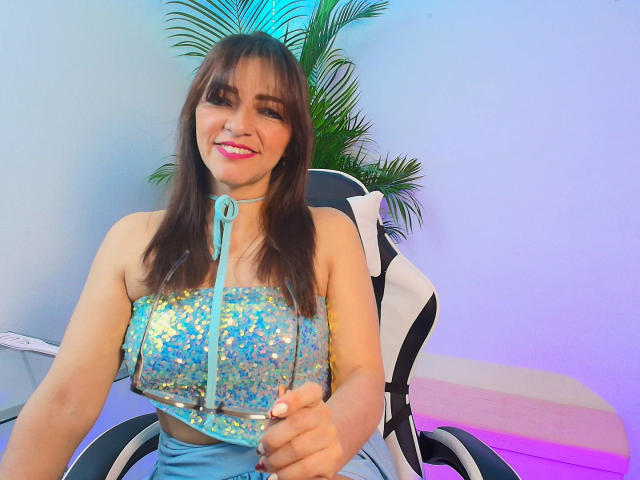 CarolMissx with sparkling blue sequin top, showing her tits, sitting in a chair, holding a leash, smiling with pink lipstick, brown hair, and green plant in the background.