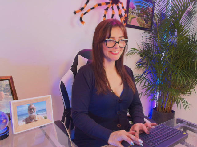 CarolMissx com óculos, cabelo castanho e seios, vestindo um cardigã azul marinho, digitando em um teclado, sorrindo e usando uma tiara de aranha em um quarto aconchegante com uma planta..