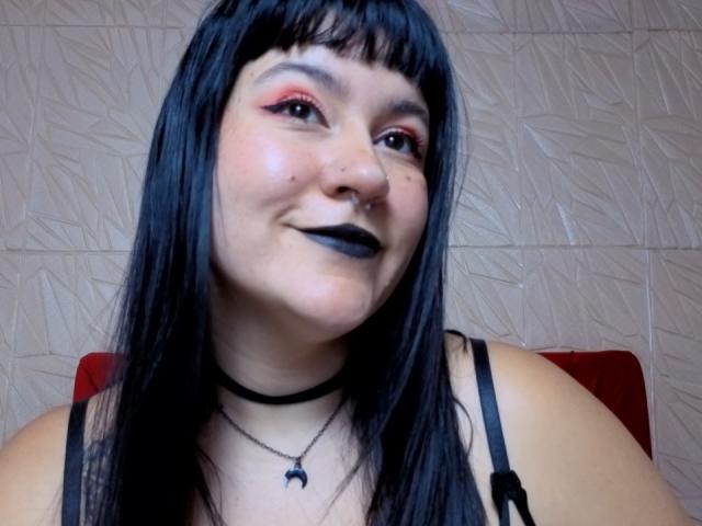 NaniSky has black hair, a nose ring, and black lipstick. She's smiling with red eyeshadow, wearing a black choker and a necklace with a small crescent moon.