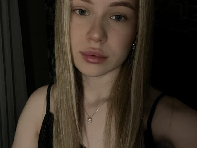 Le visage de DarceyJarver, avec ses cheveux blonds lisses, ses yeux bleus et ses lèvres roses, portant un haut noir et un petit collier avec un pendentif, dans un environnement peu éclairé..