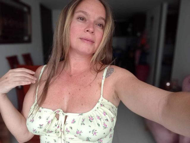 PamelaColin with light brown hair, wearing a floral, white, spaghetti-strap top, showing her small tits and a tattoo on her right arm, taking a selfie.