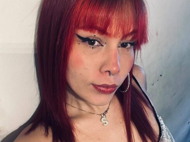 LiaLuxx tem cabelo ruivo, delineador preto, um piercing no nariz, brincos de argola e um colar com um pingente em formato de "S", vestindo uma blusa preta de renda, mostrando um rosto levemente corado..