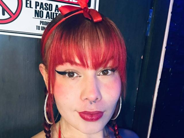 LiaLuxx tem cabelo ruivo com franja, um laço vermelho, brincos de argola, delineador preto, batom vermelho, um piercing no nariz e um piercing no septo, olhando para a câmera com um leve sorriso..