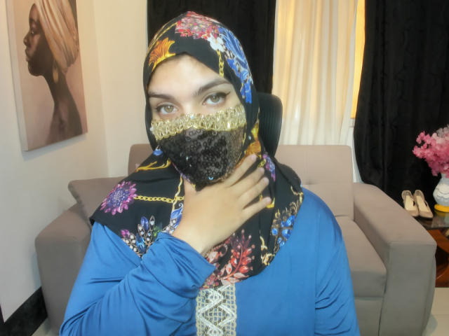 AniisaAli with floral hijab, gold lace mask, blue top, hand near neck, light brown eyes, medium tits, tan skin, black and colorful patterned scarf, gray sofa in background.