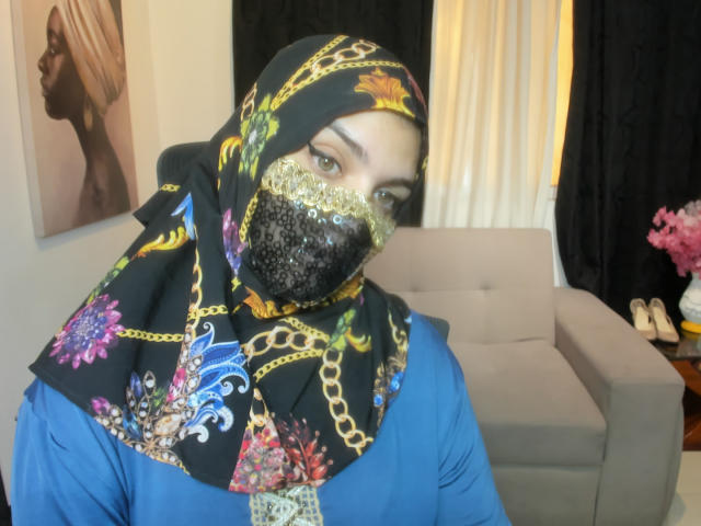 AniisaAli wearing a black hijab with colorful floral and chain patterns, blue dress, and glittery black mask covering her mouth and nose. Her eyes are looking forward.