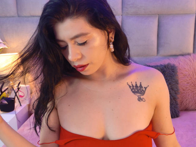 LindaMancini with long dark hair, red lipstick, and a crown tattoo on her right boob, wearing an off-shoulder red top, touching her hair, in a softly lit room.
