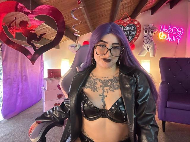 LilithDark69 met paars haar, een zwarte leren bustier, bezaaid met studs, en een leren jas, een bril, tatoeages, zwarte lippenstift, zittend in een kamer met neonborden en hartdecoraties..