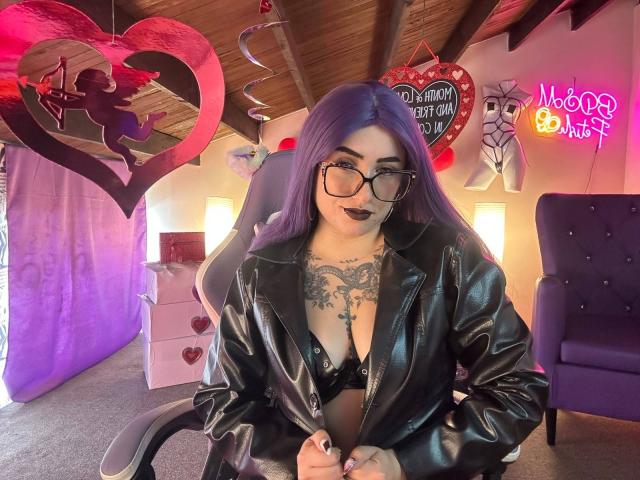 LilithDark69 with purple hair, glasses, black leather jacket, black bra, chest tattoo, dark lipstick, sitting in chair, wooden ceiling, purple curtain, pink neon sign, and heart decor.