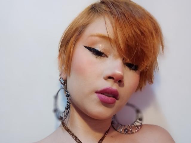 ChloeeWilliams has short orange hair, bold black eyeliner, pink lips, and wears large chain earrings. Her eyes are closed, and she's wearing a gold necklace.