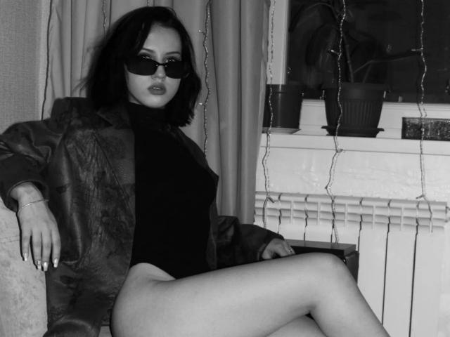 Black-and-white photo of JannaCorsaro with dark hair, sunglasses, leather jacket, black turtleneck, and no pants, showing her ass and crossed legs, sitting by a window with string lights.
