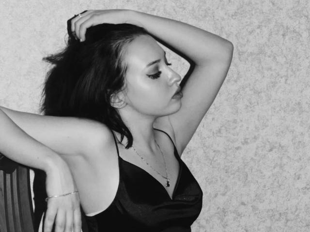 JannaCorsaro with dark hair, eyes closed, wearing a black satin dress, arm raised, hand in hair, sitting, subtle makeup, necklace, textured wallpaper background, relaxed, sexy.