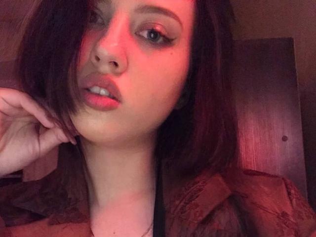 JannaCorsaro with red-lit face, medium brown hair, and dark eyeliner, wearing a brown shirt, hand by her chin, looking at the camera with slightly parted lips.