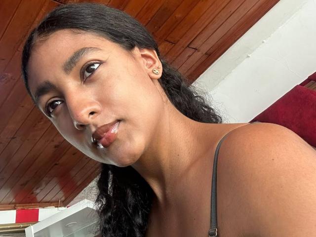NatalyBlonder has medium brown skin, black wavy hair, small earrings, and is smiling slightly with shiny red lips. She's wearing a grey spaghetti strap top.