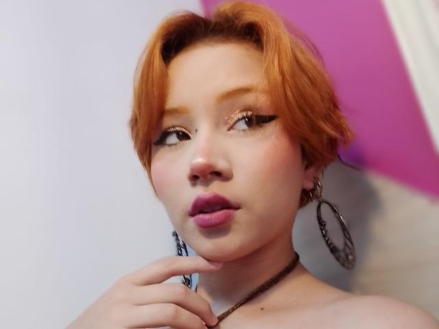 ChloeeWilliams with short orange hair, looking sideways, wearing pink lipstick, thick black eyeliner, large hoop earrings, and a black choker, touching her chin with her right hand.