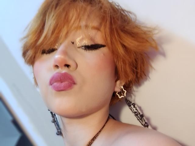 ChloeeWilliams with short orange hair, glossy pink lips, black winged eyeliner, star earrings, chain necklace, pouty face, and shiny gold eyeshadow.