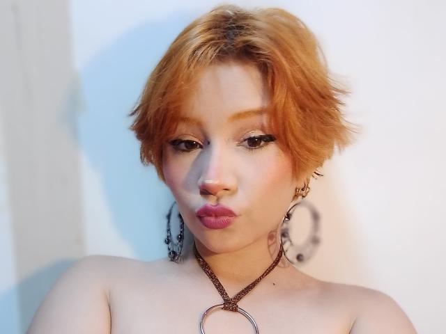 ChloeeWilliams with short orange hair, pouty pink lips, wearing a necklace with a large ring, and hoop earrings, looking directly at the camera. Her tits are partially visible.