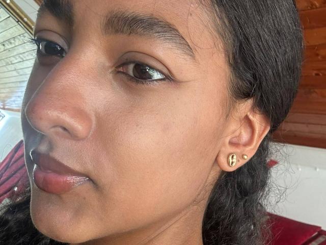 Il viso di NatalyBlonder, che mostra la sua pelle scura, i capelli neri e gli occhi marroni, con orecchini a forma di cuore dorati e labbra leggermente schiuse di colore rosato. Il suo naso è piccolo e guarda di lato..