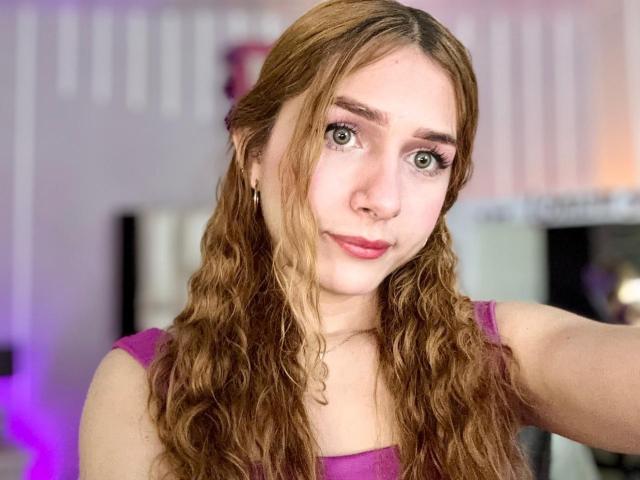 AndyDupont has wavy brown hair, green eyes, pink lipstick, and wears a purple top. Her tits are small, and she's giving a cute, subtle smile.