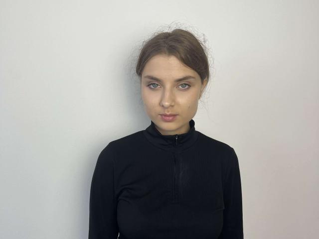 NatalieBasey has blue eyes, a nose ring, and brown hair in a messy bun. She's wearing a black, long-sleeved zip-up top, looking serious with a slight pout.