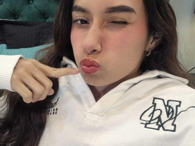 MiliBlom with pink pout, winking, her finger touching her lower lip, wearing a white hoodie with a black "BV" logo, her brown hair falling over her shoulders.