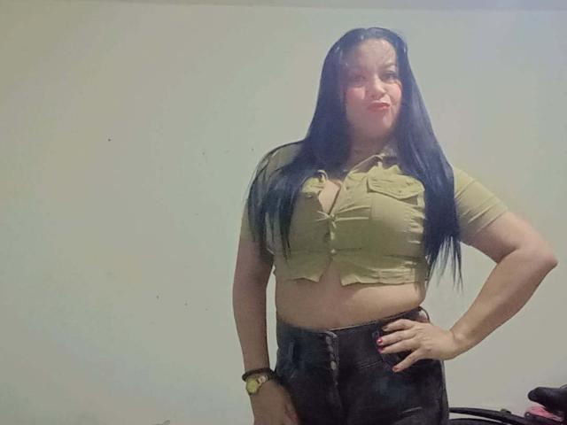 VictoriaLands with long black hair, tan crop top showing her tits, black jeans, red nails, and a watch, standing with one hand on her hip, smiling.