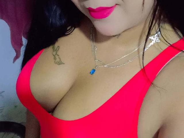 VictoriaLands in a bright red tank top with big tits, pink lipstick, blue gem necklace, and a rabbit tattoo on her right boob. Her face is partially visible.