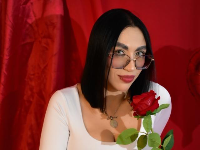 CamilleJuicyHot with black hair, glasses, white top, red rose, and necklace, smiling at camera, holding rose in front of red backdrop. Boobs visible under top.