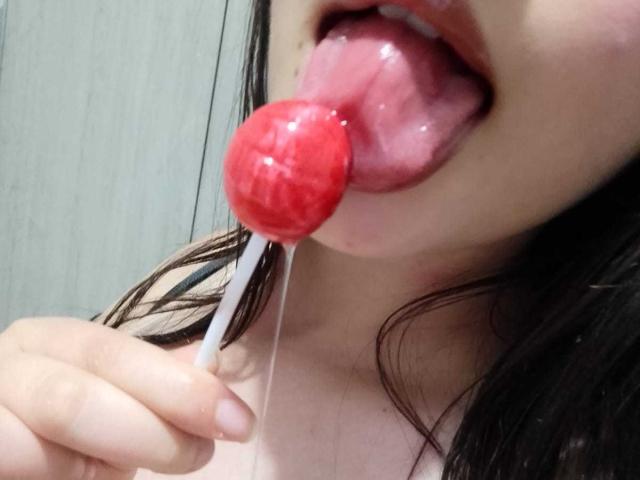 AmberBelle's pink tongue is out, licking a shiny red lollipop with a white stick, drooling slightly, showing just her lower face and hand.
