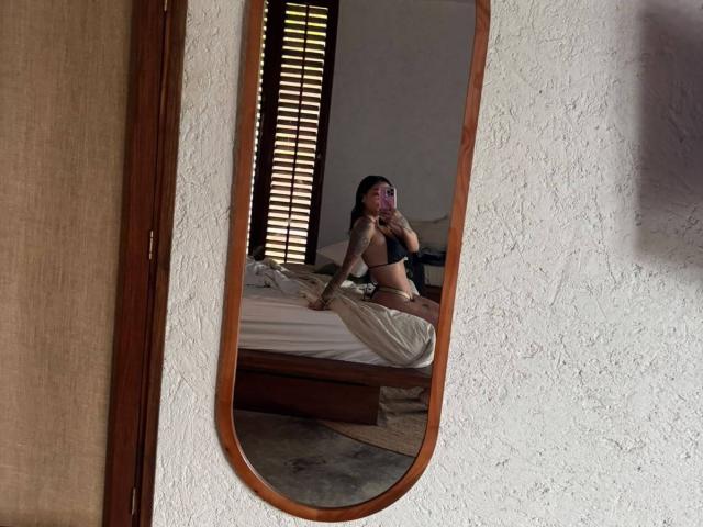 MajoJones with tits in black lingerie, sitting on a bed, reflected in an oval mirror, holding a phone, white textured wall, wooden window blinds, and bed with beige sheets.