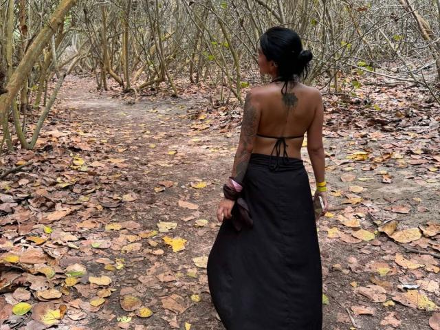 MajoJones has a tattoo on her back, wearing a black halter top and skirt, holding a brown bag, with visible tattoos on her arm, in a leafy forest path.