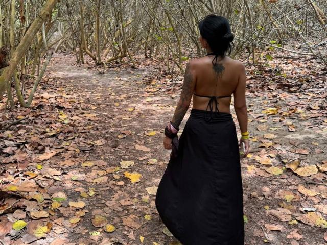 MajoJones with black hair, back tattoo, brown topless, black skirt, yellow bracelets, walking on leaf-covered forest path, autumn leaves on ground, looking ahead.
