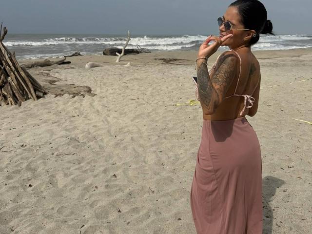 MajoJones with tattoos, wearing a pink backless dress, standing on a sandy beach, looking over her shoulder, touching her lips with fingers, wearing sunglasses.