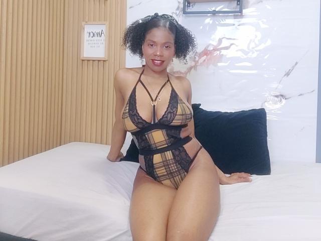 SarahiPusey with curly hair, wearing a plaid lace-up lingerie, sitting on a bed, showing her big ass and big boobs, looking at the camera with a smile.