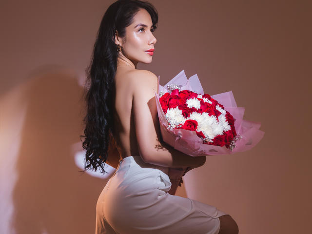 ArianaRogers with long black hair, topless, holding a bouquet of red and white flowers, wearing white pants, looking over her shoulder, showing her ass and back.