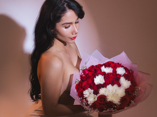 ArianaRogers with long black hair, holding a red and white rose bouquet, topless, showing her tits, wearing a necklace, in a softly lit, brown-hued setting.
