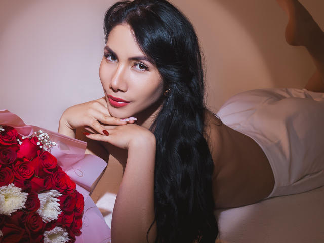ArianaRogers with long black hair, lying on a bed, wearing white lingerie, holding a red rose bouquet, looking at the camera with a sexy smile, red nails.