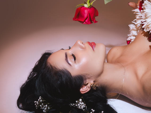 ArianaRogers with dark hair, glossy skin, and red lipstick, lying down, eyes closed, holding a red rose above her face, wearing a white flower in her hair.