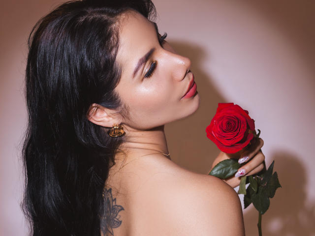 ArianaRogers with long black hair, holding a red rose to her nose, her bare shoulders showing a tattoo, wearing gold earrings and a necklace, with a sexy smile.