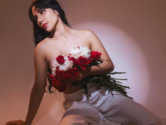 ArianaRogers with dark hair, red lipstick, holding red roses and white chrysanthemums, wearing white pants, sitting, light shadow on her, soft light, looking to the side.