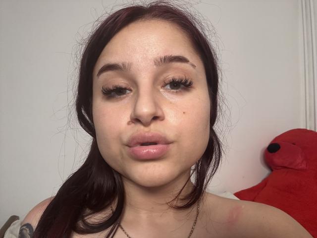 Luucie hat helle Haut, dunkelbraunes Haar im Pferdeschwanz, dicke Augenbrauen, lange Wimpern und glänzlich rosafarbene Lippen. Ein rotes Stofftier ist rechts zu sehen. Ein kleines Tattoo befindet sich auf der linken Schulter..