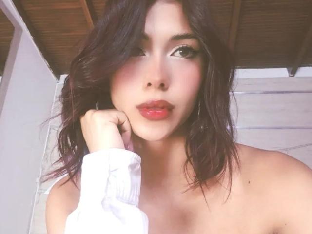 DanikaSerov with wavy dark hair, red lipstick, and white shirt sleeve, touching her cheek, showing bare shoulders, looking at the camera with slightly parted pink lips.