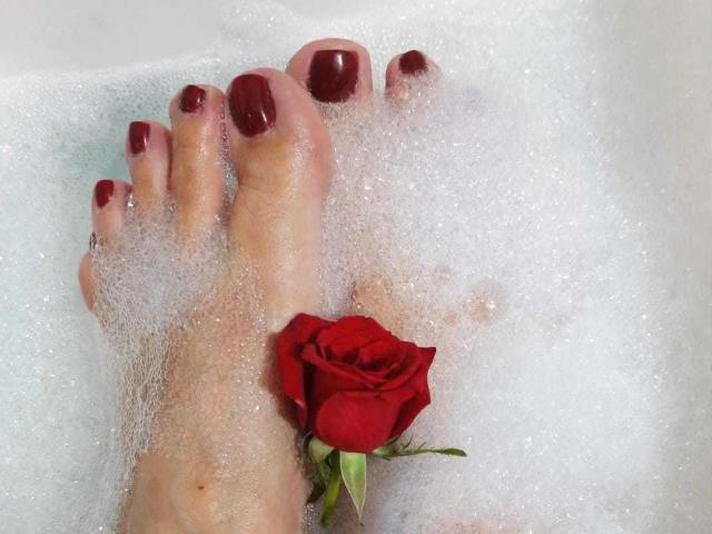 VanesaMokas Füße in einem seifigen Bad, mit rot lackierten Zehennägeln, eine einzelne rote Rose zwischen ihren Zehen und weißem Schaum, der ihre Haut bedeckt..