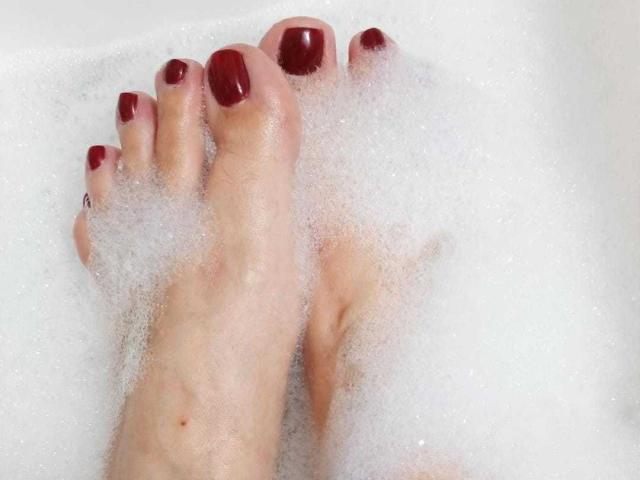 VanesaMoka's feet, with dark red painted toenails, partially covered in white bubble foam, showing a small tattoo on the big toe.