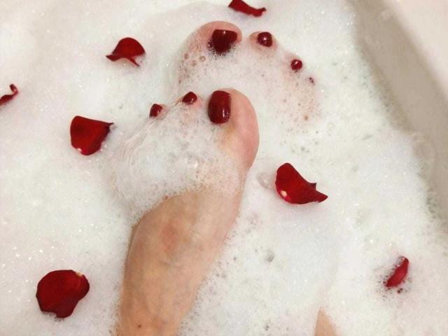 VanesaMoka's foot, with painted red toenails, partially submerged in a white, foamy bath, surrounded by scattered red rose petals.