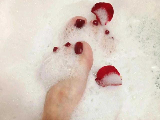 O pé de VanesaMoka, com esmalte vermelho escuro, parcialmente submerso em espuma de banho branca, com pétalas de rosa vermelhas flutuando ao redor..