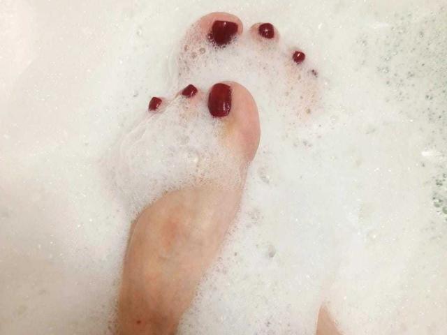 VanesaMoka's feet, with red-painted toenails, partially submerged in white, foamy bath water. Her toes are visible through the soap bubbles.