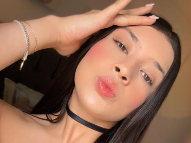 EmmaLombardi with black choker, pouty pink lips, long black hair, brown eyes, and a gold bracelet on her left hand, touching her head. She's looking at the camera.