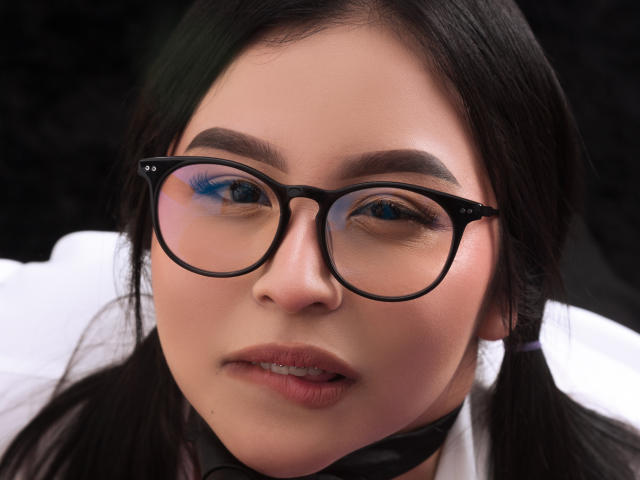 AlaiaSmile has black puffy-rimmed glasses, blue eyeshadow, and puffy pink lips. She's wearing a black choker with white shirt. Her dark hair is in pigtails.