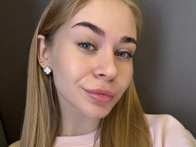 BarbaraBlum has blonde hair, blue eyes, and pink lips with a slight smile. She's wearing a pink top and flower earrings. Her skin is fair and smooth.